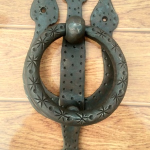May include: A dark metal door knocker with a ring handle and a trident-shaped backplate. The ring is decorated with star-shaped designs. The backplate has a series of small holes and is mounted on a wooden door.