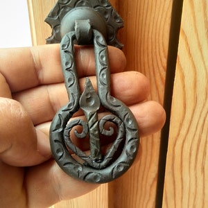May include: A black metal door knocker with a decorative design. The knocker is shaped like a heart with a swirl pattern and is attached to a triangular base.
