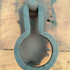 May include: A rusty, black metal door knocker with an ornate design. The knocker is shaped like a teardrop with a ring handle.