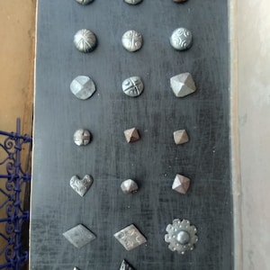 May include: A collection of 19 different metal nail heads with various shapes and designs. The nail heads are arranged in rows on a dark grey surface.