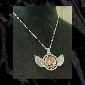 May include: Silver chain necklace with a round pendant featuring a photo of a man with dreadlocks. The pendant is surrounded by clear rhinestones and has two wings on either side.
