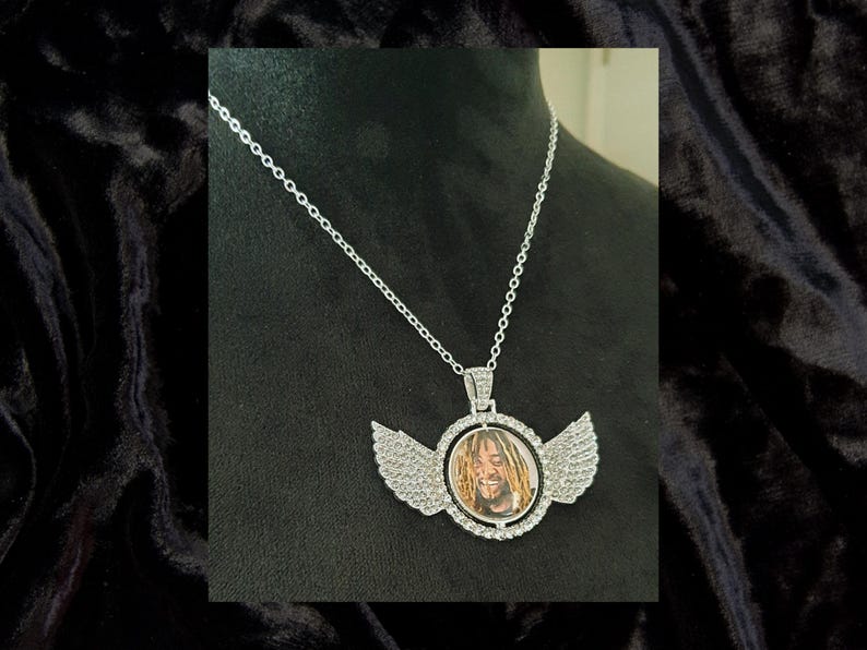 Puede incluir: Collar de cadena de plata con un colgante redondo que presenta una foto de un hombre con rastas. El colgante est&aacute; rodeado de diamantes de imitaci&oacute;n transparentes y tiene dos alas a cada lado.