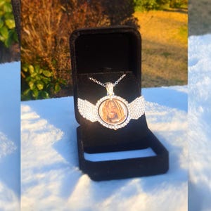 Puede incluir: Un collar con un colgante redondo de plata con una foto de una persona con rastas. El colgante est&aacute; rodeado de diamantes de imitaci&oacute;n transparentes y tiene dos alas a cada lado. El collar est&aacute; en una caja de terciopelo negro.