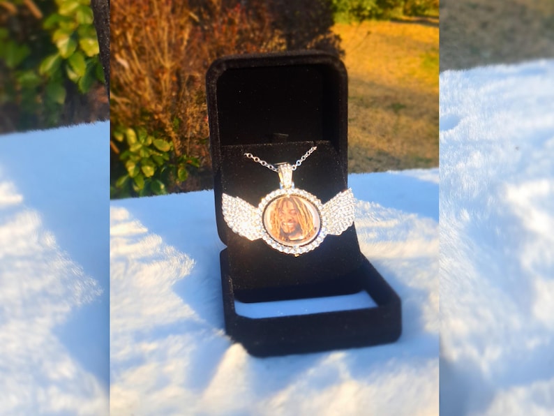 Puede incluir: Un collar con un colgante redondo de plata con una foto de una persona con rastas. El colgante est&aacute; rodeado de diamantes de imitaci&oacute;n transparentes y tiene dos alas a cada lado. El collar est&aacute; en una caja de terciopelo negro.