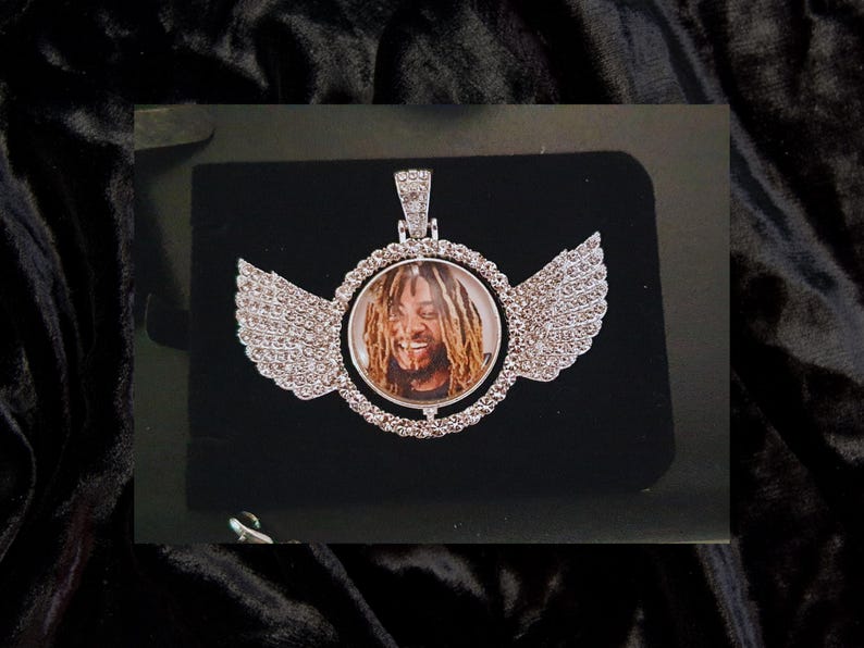 Puede incluir: Un colgante de plata con una foto redonda transparente de un hombre con rastas. El colgante est&aacute; rodeado de piedras de cristal transparentes y tiene dos alas a cada lado, tambi&eacute;n cubiertas de piedras de cristal.