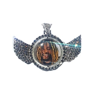 Puede incluir: Un colgante de plata con un inserto de foto redondo transparente que presenta a un hombre con rastas. El colgante est&aacute; rodeado por un halo de peque&ntilde;as piedras plateadas y tiene dos alas a cada lado, tambi&eacute;n adornadas con piedras plateadas.