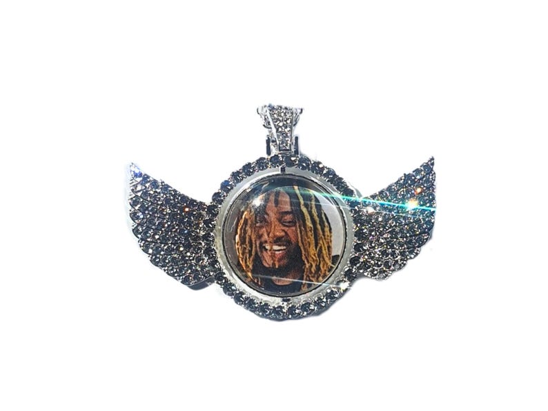 Puede incluir: Un colgante de plata con un inserto de foto redondo transparente que presenta a un hombre con rastas. El colgante est&aacute; rodeado por un halo de peque&ntilde;as piedras plateadas y tiene dos alas a cada lado, tambi&eacute;n adornadas con piedras plateadas.