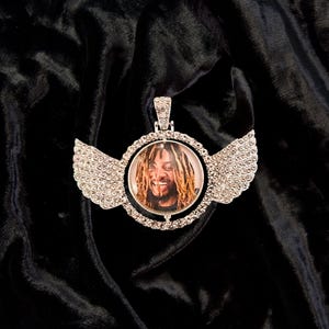 Puede incluir: Un colgante de plata con un marco redondo, transparente, incrustado de cristales, que presenta una foto de un hombre con rastas. El colgante tiene dos alas de plata con detalles de cristal.