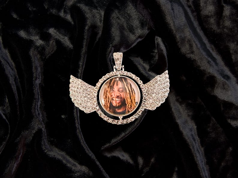 Puede incluir: Un colgante de plata con un marco redondo, transparente, incrustado de cristales, que presenta una foto de un hombre con rastas. El colgante tiene dos alas de plata con detalles de cristal.