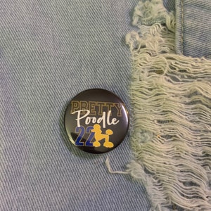 May include: A black button with gold lettering that says "Pretty Poodle 22" with a gold silhouette of a poodle in the center.
