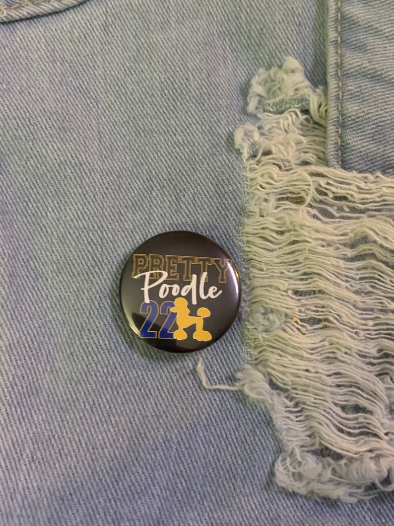 May include: A black button with gold lettering that says "Pretty Poodle 22" with a gold silhouette of a poodle in the center.