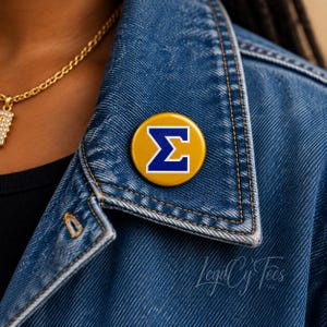 May include: A circular pinback button featuring a mustard yellow background with a bold, royal blue Greek letter Sigma symbol centered in the middle. The button is attached to the collar of a blue denim jacket with visible stitching.