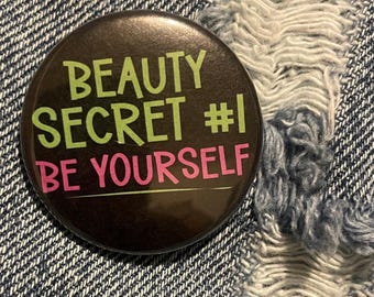 Pode incluir: Um botão preto com o texto "BEAUTY SECRET #1 BE YOURSELF" em letras verdes e rosa.