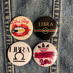 May include: Four round buttons with different designs featuring the zodiac sign Libra. One button has a red lip with a lipstick tube, another has a black background with a gold Libra symbol, another has a white background with a black Libra symbol, and the last button has a white background with a pink and black design that says "a-badass Libra".
