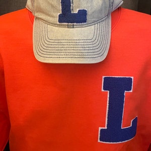 May include: An orange sweatshirt and a light gray baseball cap, both featuring a large, dark blue letter "L". The cap has a curved brim and a matching blue stitched outline. The sweatshirt has the letter patch on the chest.