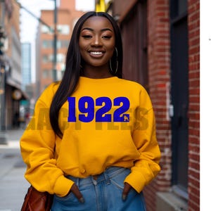 May include: A woman wearing a yellow sweatshirt with the blue number "1922" printed on the front. The sweatshirt has long sleeves and is paired with blue jeans. The background includes a city street and brick building.