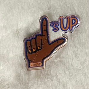 May include: A brown hand with a blue outline making the "L" hand gesture. The hand is holding a clear acrylic keychain with the text "Langston University" and the words "It's Up" in orange and blue.