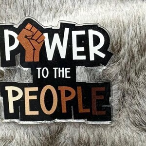 Può includere: Un cartello bianco e nero con il testo "POWER TO THE PEOPLE" in lettere bianche. Un pugno alzato è raffigurato in marrone e nero.
