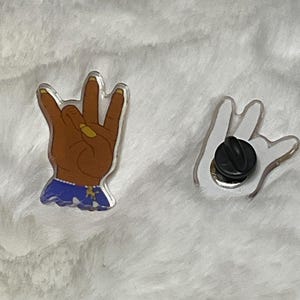 May include: Two pins, one with a hand making the 'W' sign and the other with a hand making the 'rock on' sign. The 'W' hand is brown with yellow fingernails and is wearing a blue and white patterned shirt. The 'rock on' hand is white.