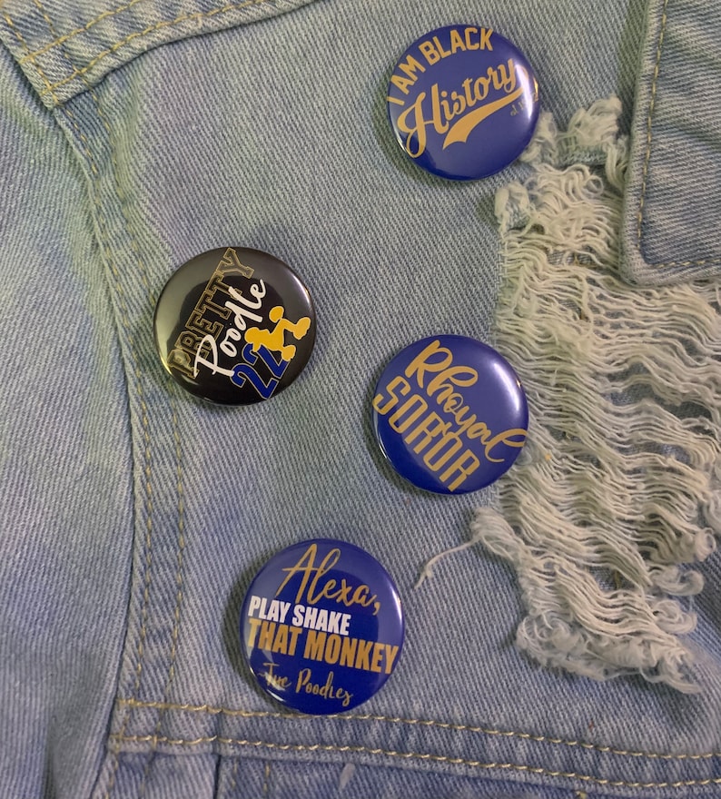 May include: Four blue and gold button pins with different slogans. The pins are on a blue denim background. The slogans are "I Am Black History", "Pretty Poodle 22", "Royal Soror", and "Alexa, Play Shake That Monkey Tue Poodies".