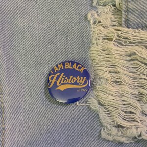 May include: A blue button with the text "I AM BLACK History est. 1922" in gold lettering. The button is on a light blue denim background.