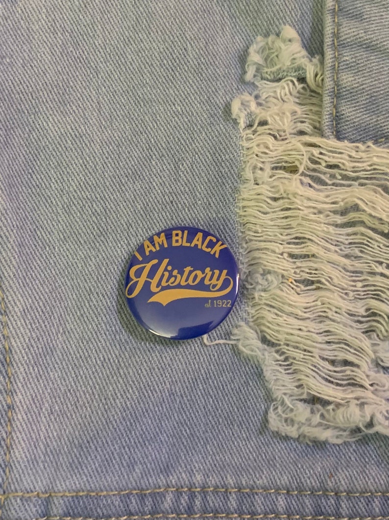 May include: A blue button with the text "I AM BLACK History est. 1922" in gold lettering. The button is on a light blue denim background.