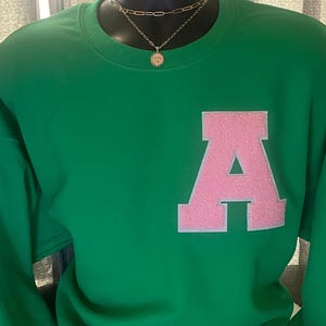 May include: Green sweatshirt with a pink felt letter A on the front. The sweatshirt has a crew neck and long sleeves.