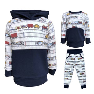 May include: A three-piece toddler boy's clothing set featuring a navy blue hooded sweatshirt, a navy blue long-sleeve shirt, and navy blue sweatpants. All pieces feature a white background with a repeating pattern of colorful trains and railroad tracks.