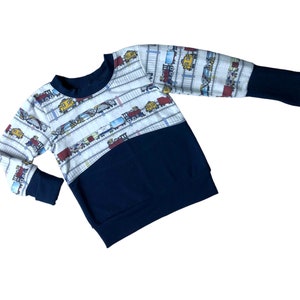 May include: A white long-sleeved sweatshirt with a navy blue bottom half. The top half features a repeating pattern of colorful toy trains on a white background.