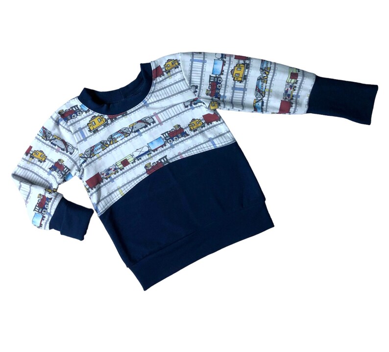 May include: A white long-sleeved sweatshirt with a navy blue bottom half. The top half features a repeating pattern of colorful toy trains on a white background.