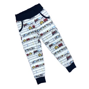 May include: White sweatpants with a navy blue waistband and cuffs. The sweatpants have a repeating pattern of colorful cartoon trains.