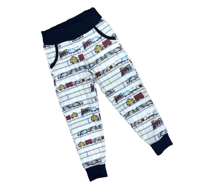 May include: White sweatpants with a navy blue waistband and cuffs. The sweatpants have a repeating pattern of colorful cartoon trains.