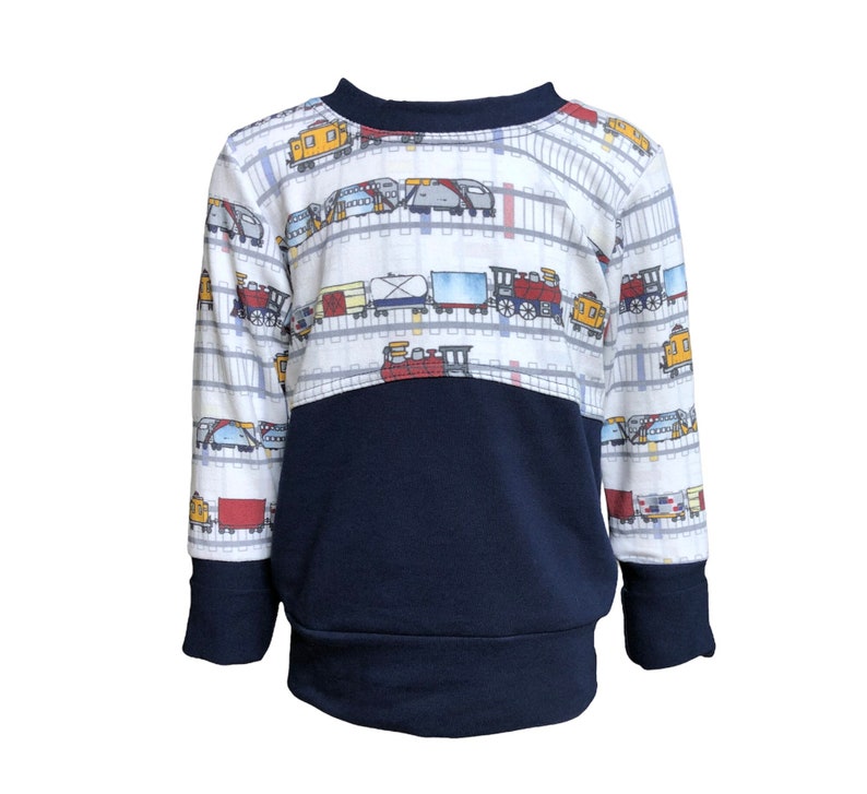May include: A white long-sleeved sweatshirt with a navy blue bottom half. The top half features a repeating pattern of colorful trains on white railroad tracks.