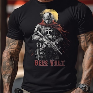 May include: Black t-shirt featuring a detailed illustration of a soldier in armor holding a rifle, with a red scarf and a yellow moon in the background. The text "DEUS VULT" is printed in red at the bottom of the design. The shirt is a crew neck.