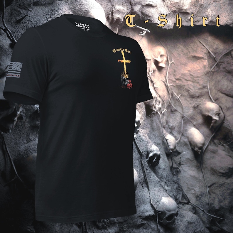 Pu&ograve; includere: T-shirt nera con la scritta "T-Shirt" in oro. La maglietta presenta un disegno a croce dorata con le parole "Memento Mori" e una piccola bandiera americana sulla manica. La maglietta &egrave; realizzata in materiale morbido.