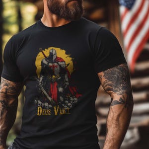 May include: Black t-shirt with a graphic of a knight in armor holding a sword and standing on a pile of skulls. The knight is wearing a red cape and a helmet with a cross on it. The text "Deus Vult" is printed below the graphic.