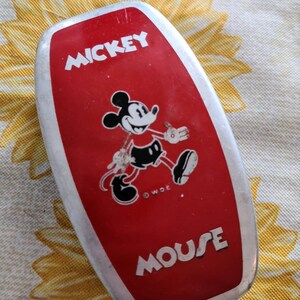 Peut inclure: Une bo&icirc;te &agrave; lunch en m&eacute;tal rouge Mickey Mouse avec une bordure argent&eacute;e. La bo&icirc;te &agrave; lunch pr&eacute;sente une image en noir et blanc de Mickey Mouse marchant et le texte "Mickey Mouse" en lettres blanches.