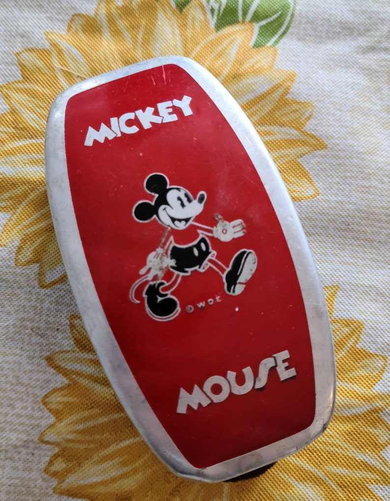 Peut inclure: Une bo&icirc;te &agrave; lunch en m&eacute;tal rouge Mickey Mouse avec une bordure argent&eacute;e. La bo&icirc;te &agrave; lunch pr&eacute;sente une image en noir et blanc de Mickey Mouse marchant et le texte "Mickey Mouse" en lettres blanches.