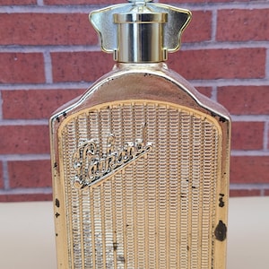 May include: A vintage gold-colored perfume bottle with a decorative design. The bottle has a rectangular shape with a detailed front resembling a car grille. The top features a circular emblem with text. The bottle is set against a brick wall.