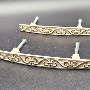 May include: Two gold-toned cabinet drawer pulls with ornate scrollwork designs. The pulls are made of metal and have a slightly aged finish.