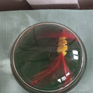 May include: A red and yellow fishing lure encased in a clear glass dome. The lure has a red feather tail and a yellow body.
