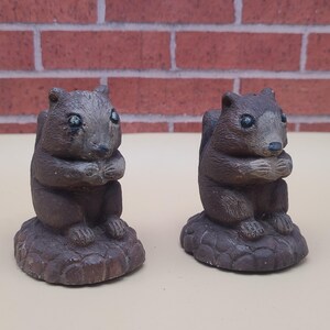 May include: Two brown squirrel figurines. Each squirrel is in a seated position, with its paws clasped together. The squirrels are on a textured, round base. The background is a brick wall.