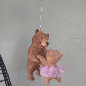 May include: A brown bear figurine dancing with a smaller bear wearing a pink tutu. The bears are holding hands, and the figurine is suspended by a silver wire. The background is a light green color.