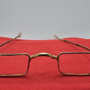 May include: A pair of vintage-style gold-toned metal spectacles featuring thin, rectangular frames and a curved bridge. The glasses have slender, straight temples that extend outward, showcasing a minimalist design with a metallic finish throughout.