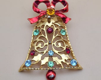 May include: A gold-tone Christmas bell brooch with a red bow and colorful rhinestones. The bell is decorated with a filigree design and has a small red ball at the bottom.