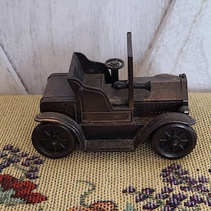 May include: A vintage-style metal pencil sharpener shaped like a car. The car is brown and has a black steering wheel and wheels. It is sitting on a patterned fabric surface.