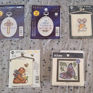 May include: Five cross-stitch kits. Two kits feature religious designs with text. One kit has a mouse with the text "Say Cheese". Another kit shows a bear with bees. The last kit shows a butterfly.