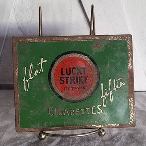 May include: Vintage Lucky Strike cigarettes tin with a green background, red and white circle logo, and the text "flat fifties" written in cursive. The tin is slightly rusted and has a worn appearance.