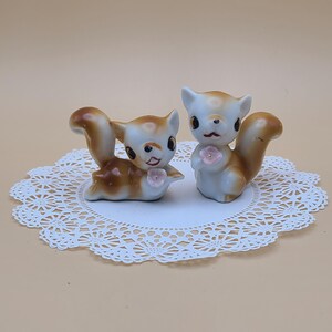 May include: Two ceramic squirrel figurines. One is lying down, the other is standing upright. Both have brown and white fur, black eyes, and pink flower accents. They are placed on a white lace doily.