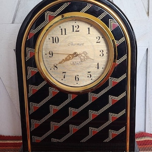 May include: A black and gold tabletop clock with a gold face and black hands. The clock has a red and gold geometric pattern on the front. The clock face reads "Charmant Quartz".